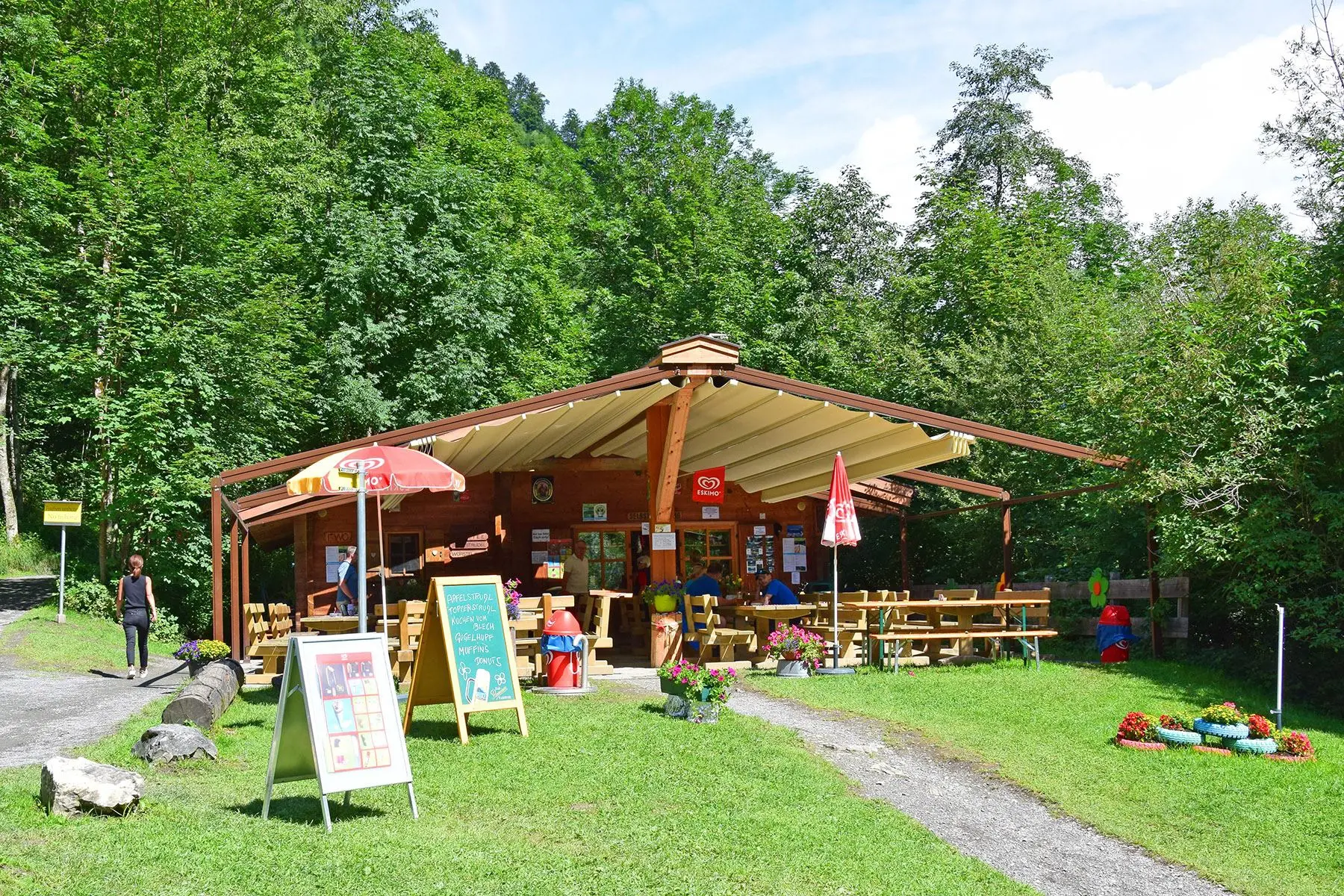We are looking head-on at the Klammseestüberl in Kaprun on a warm, sunny summer's day. A woman is walking along the hiking trail to the left. The menu boards are clearly visible in front of the parlour.