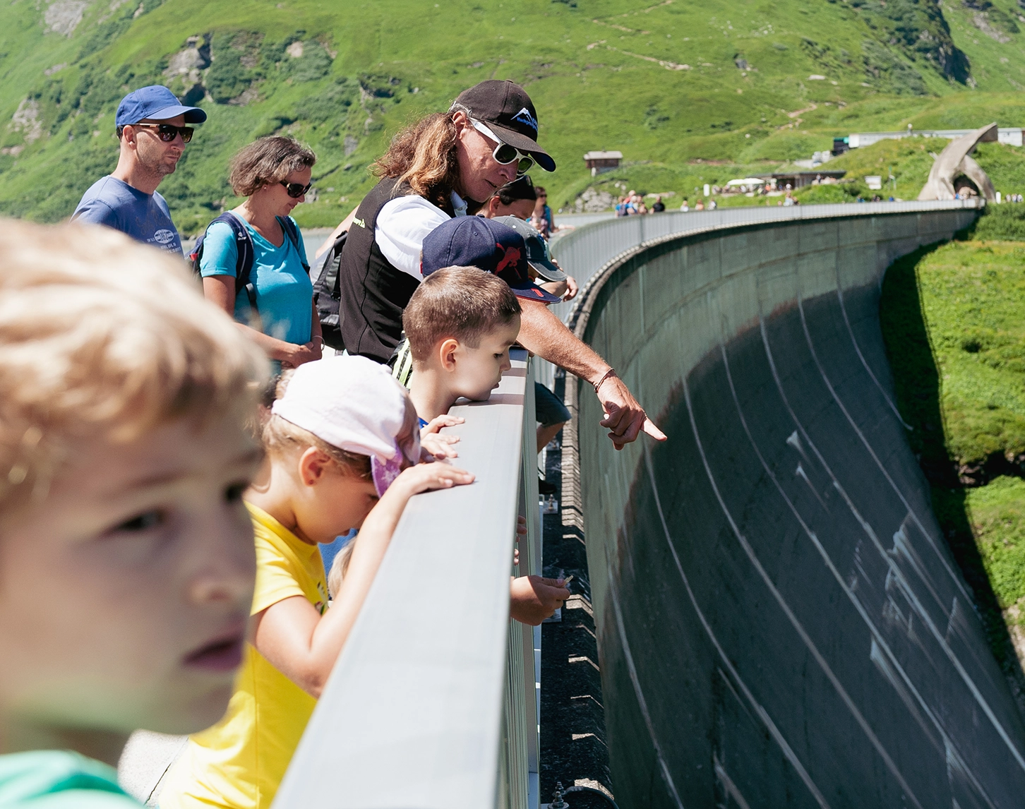 Wir blicken auf eine Kinderführung in Kaprun. Der Gruppenführer blickt mit den jungen Interessierten über die Staumauer in die Tiefe. Im Hintergrund sehen wir grüne Wiesen.