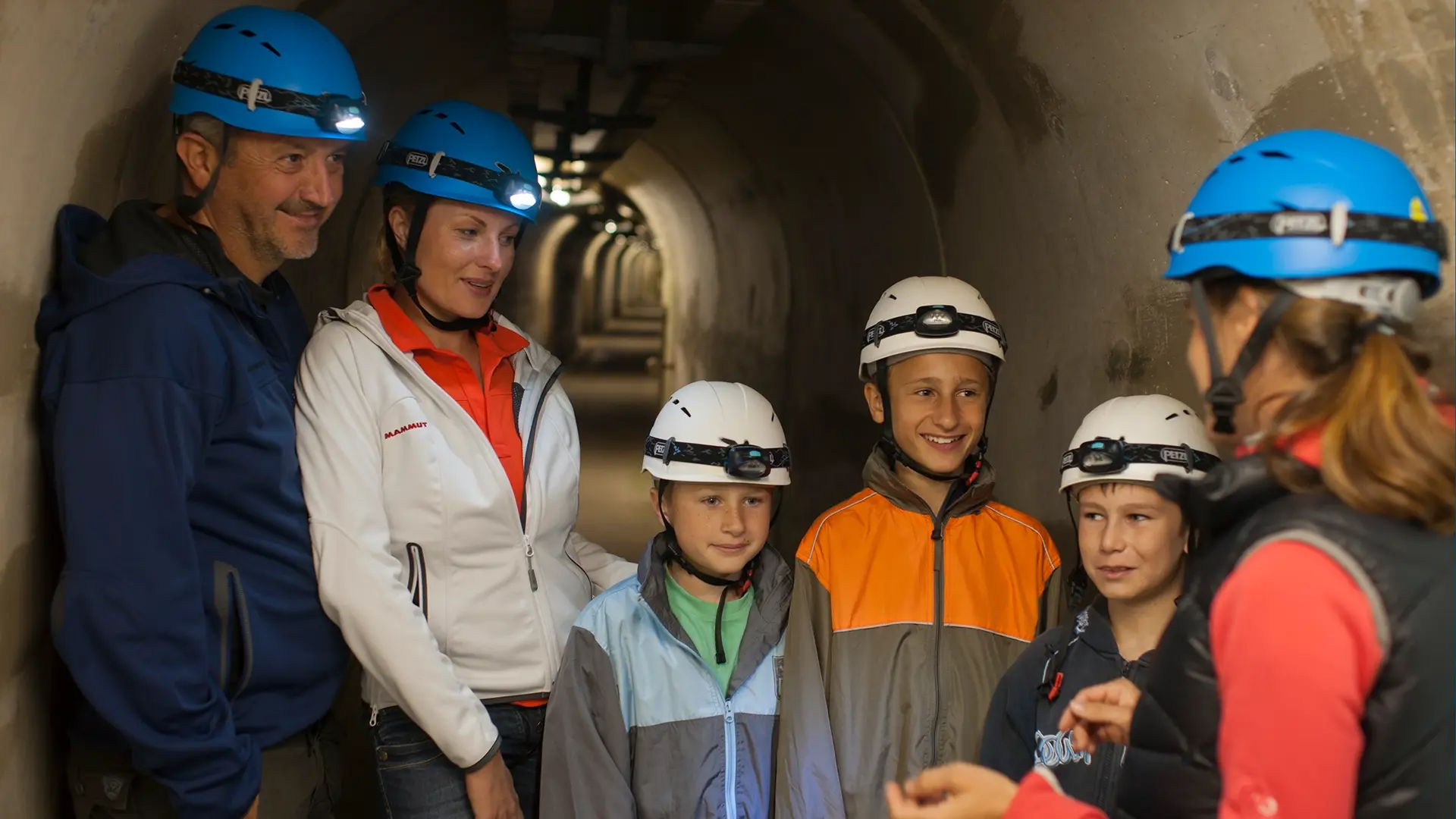 Wir sehen eine Familie bei der Staumauerführung mit einer Führerin in der Staumauerführung. Drei Kinder und zwei Eltern tragen Freizeitkleidung und alle tragen einen Schutzhelm.