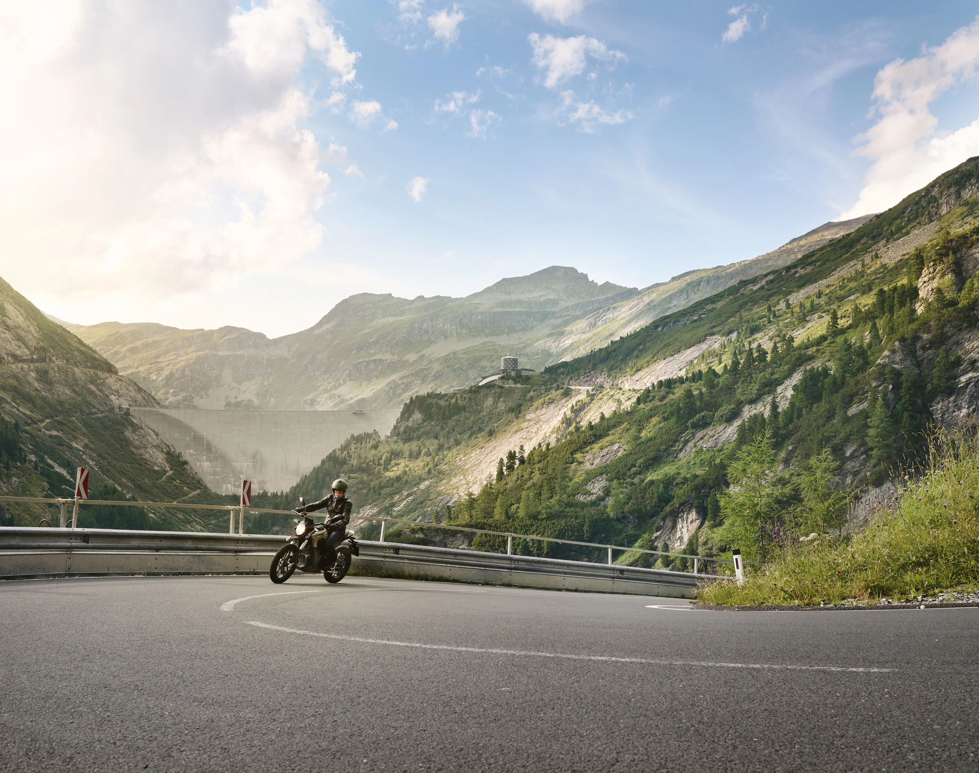 A motorcyclist rides up a mountain road to the dam in Malta. In the background, we see mountains and sunshine.