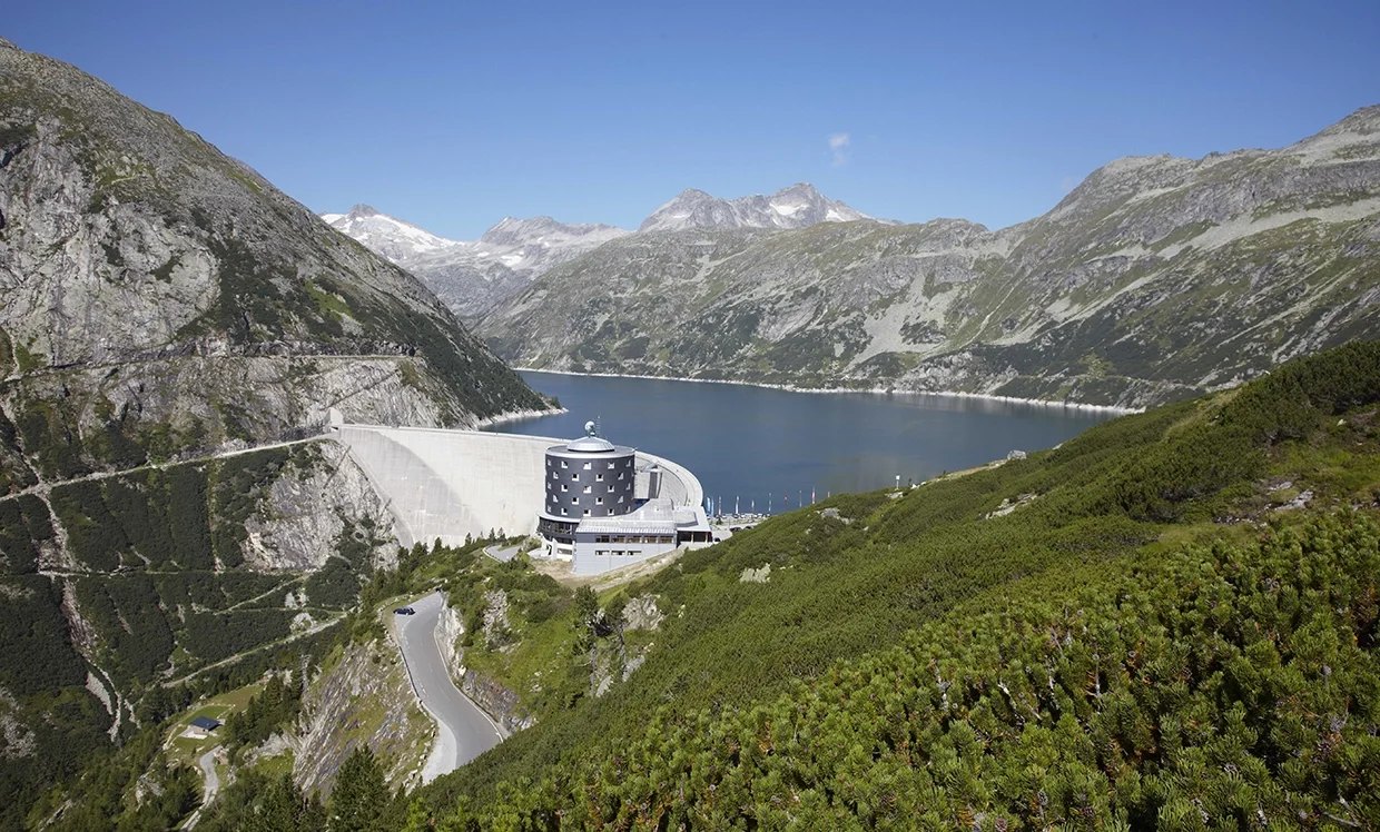 Wir blicken von einer erhöhten Position auf das Berghotel Malta. Die Sonne scheint. Der Stausee liegt in Ruhe.