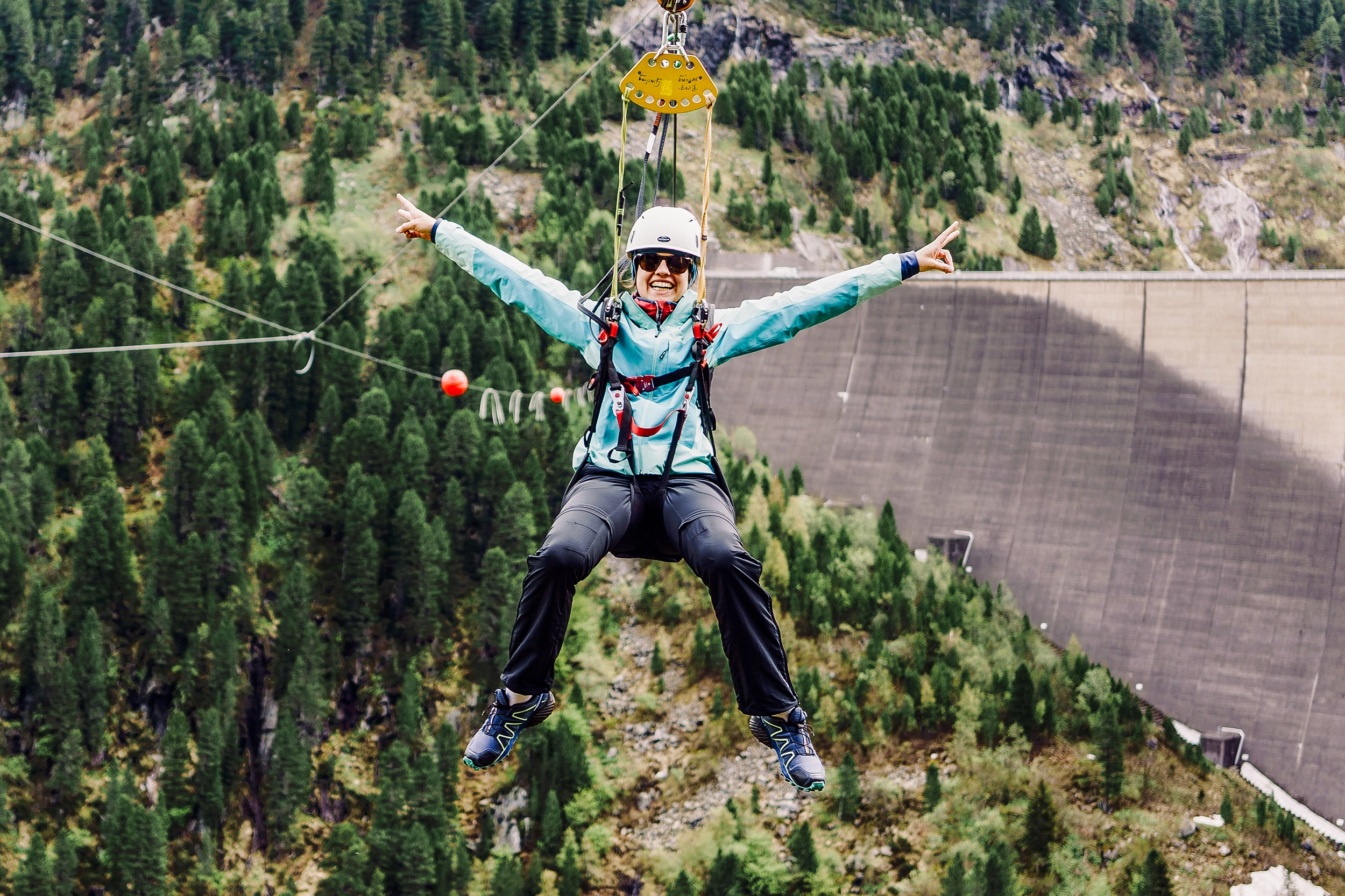 A young woman hurtles through the air on the Flying Fox in Schlegeis. We see her head-on in the air. She is wearing the necessary equipment and sunglasses. She laughs with both cheeks and has her hands up.