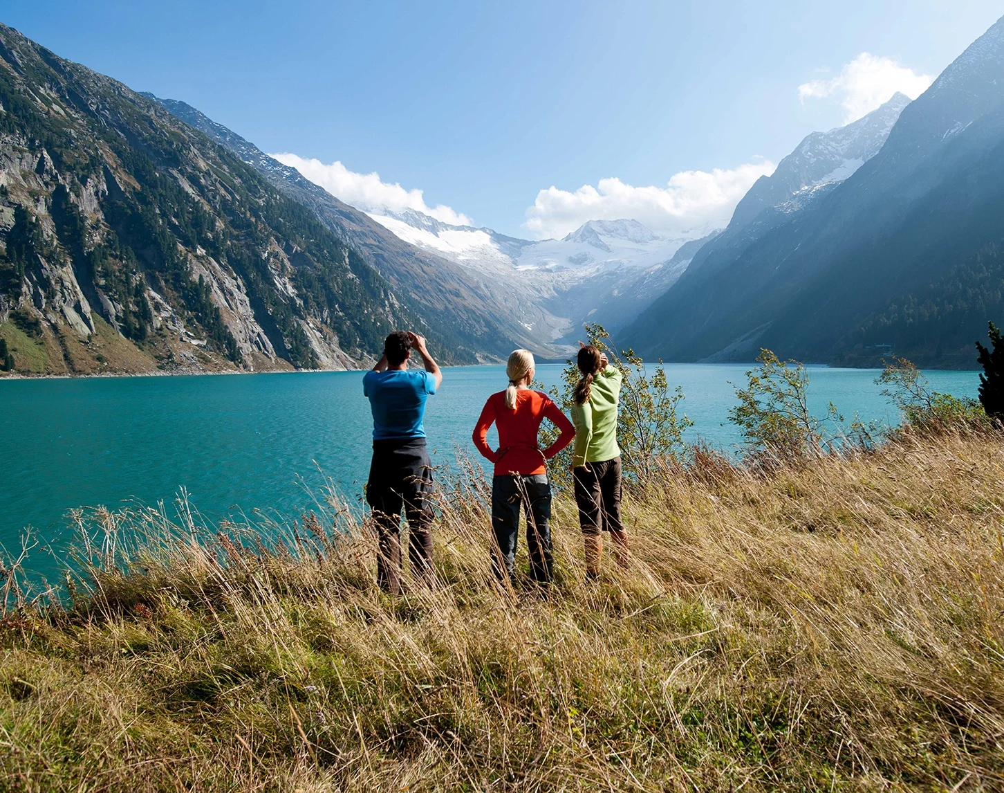 Three hikers are standing on a slightly brownish mountain meadow at the reservoir in Schlegeis. We can only see their backs. The lake and the Alps can be seen in the background. The sky is blue.