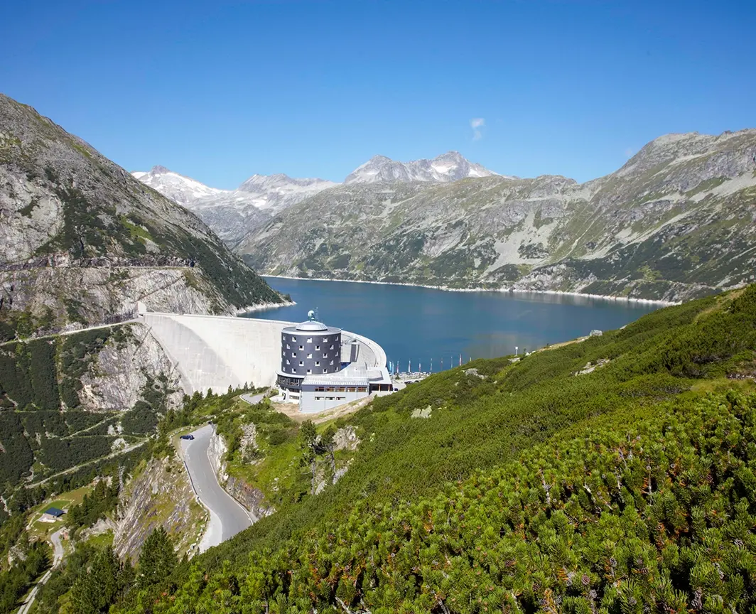Wir blicken direkt auf das Berghotel Malta. Im Vordergrund sehen wir eine Straße. Im Hintergrund sind Berge und der Stausee.