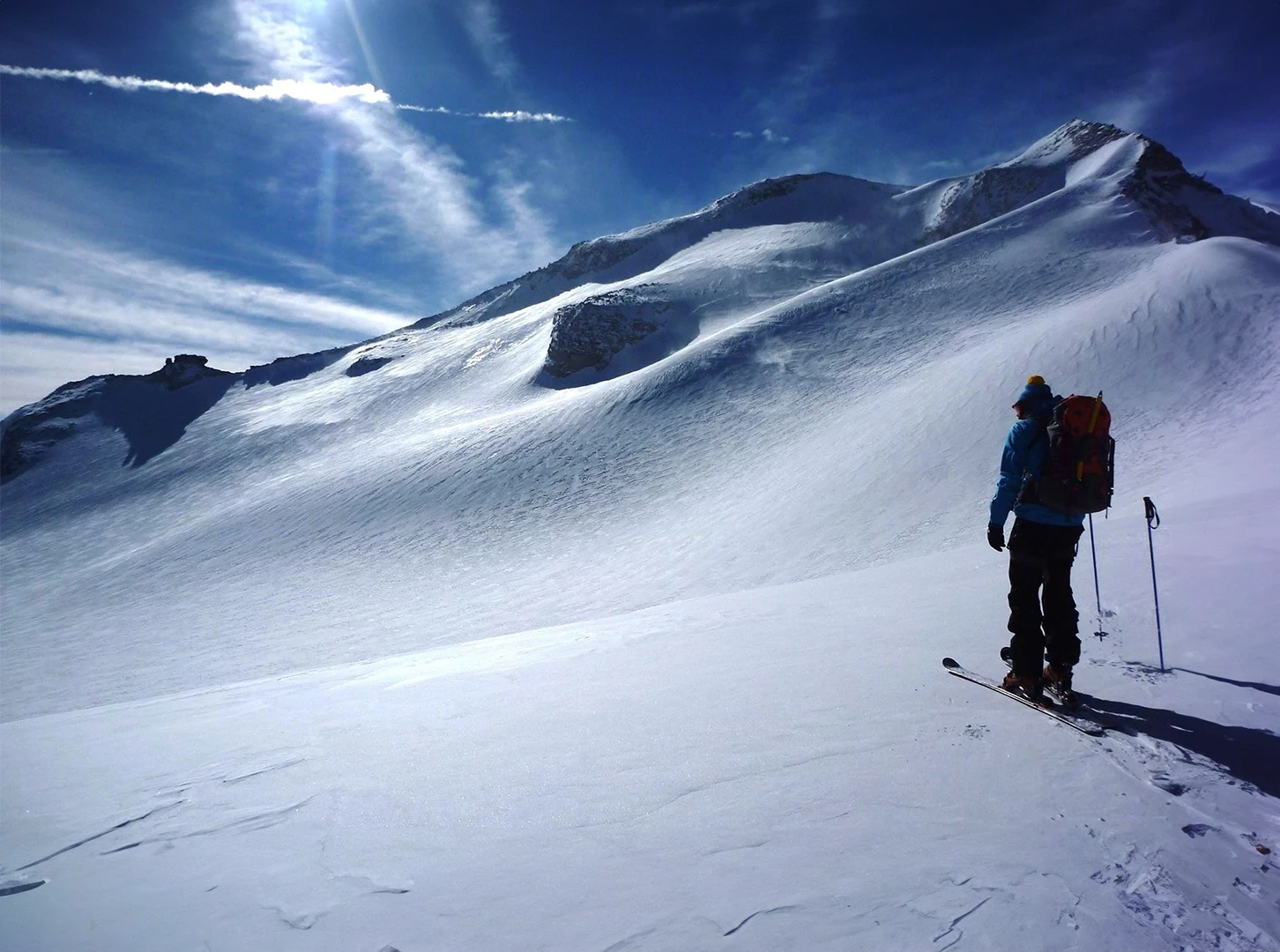 We look out over a snowfield in the middle of the high mountains. The winter sun is shining. On the right-hand side of the snowfield is a ski tourer who has put his poles aside in the snow to enjoy the view.