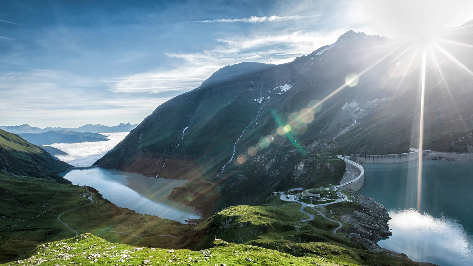 Sunrise at the Mooserboden reservoir in Kaprun: the sun peeps over the mountains, wafts of mist billow out of the depths, the sky turns blue.