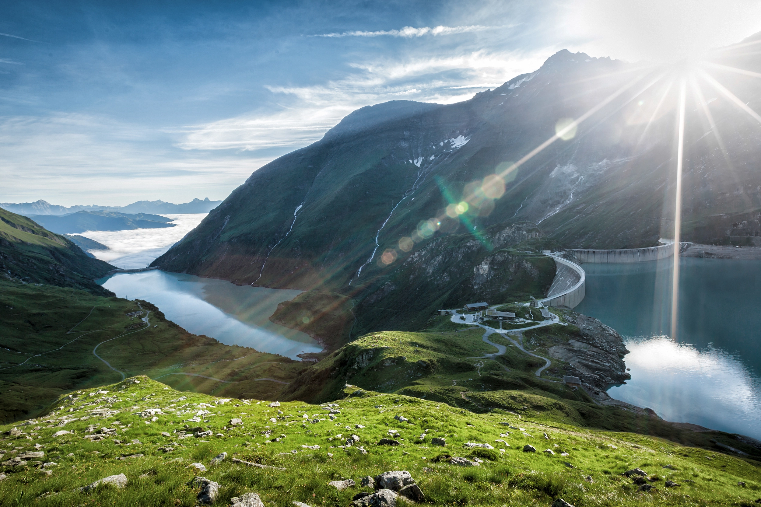 Sunrise at the Mooserboden reservoir in Kaprun: the sun peeps over the mountains, wafts of mist billow out of the depths, the sky turns blue.