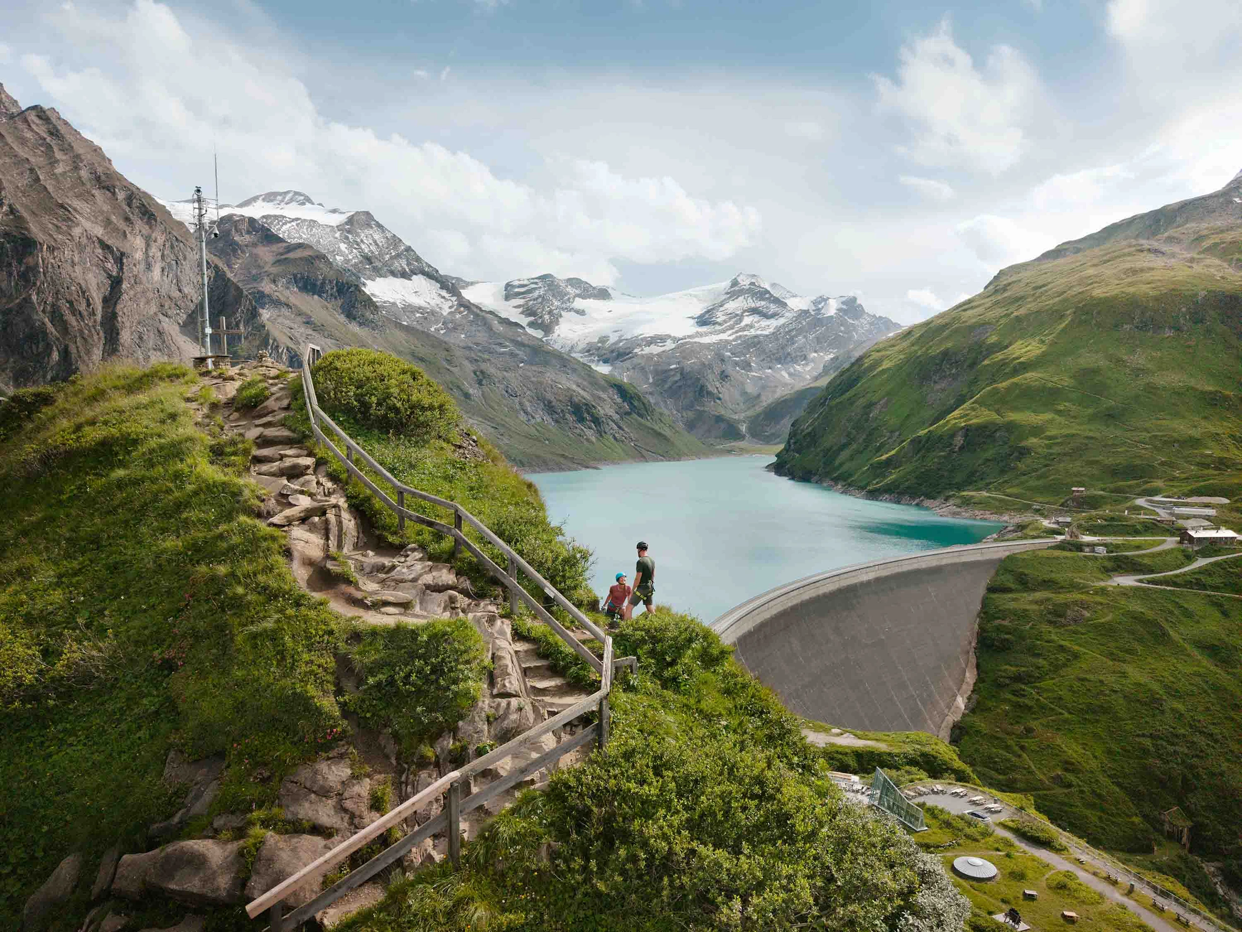 A view of the hilltop castle in Kaprun. We can see the lake in the middle ground. A couple in climbing gear are making their way up the final few metres to the summit. Mountains and clouds form the backdrop.