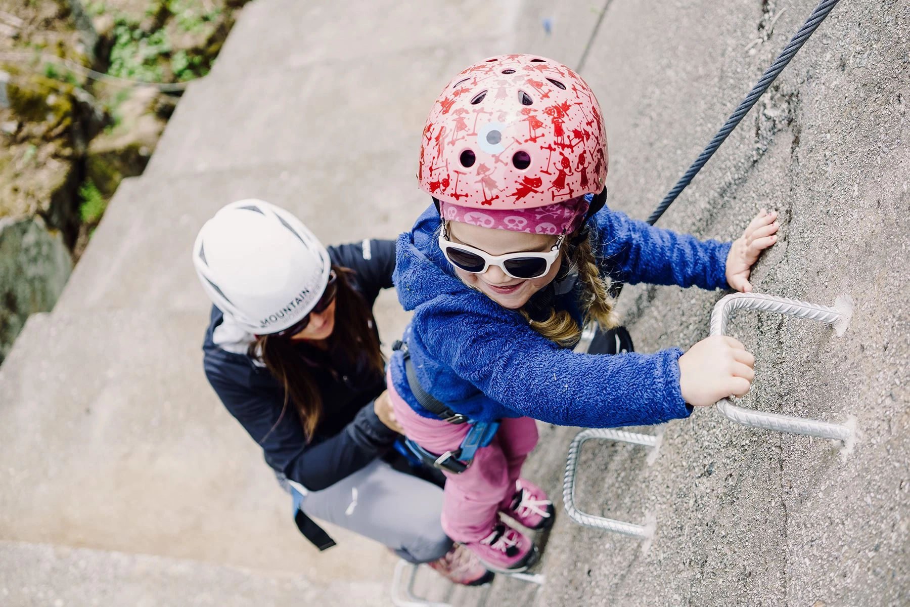 We see a child with adult company on the climbing wall in Schlegeis. The girl is wearing a red and pink patterned helmet, a blue plush waistcoat, pink trousers and pink trainers. She is secured by a harness on the climbing steps and is supported by an adult.