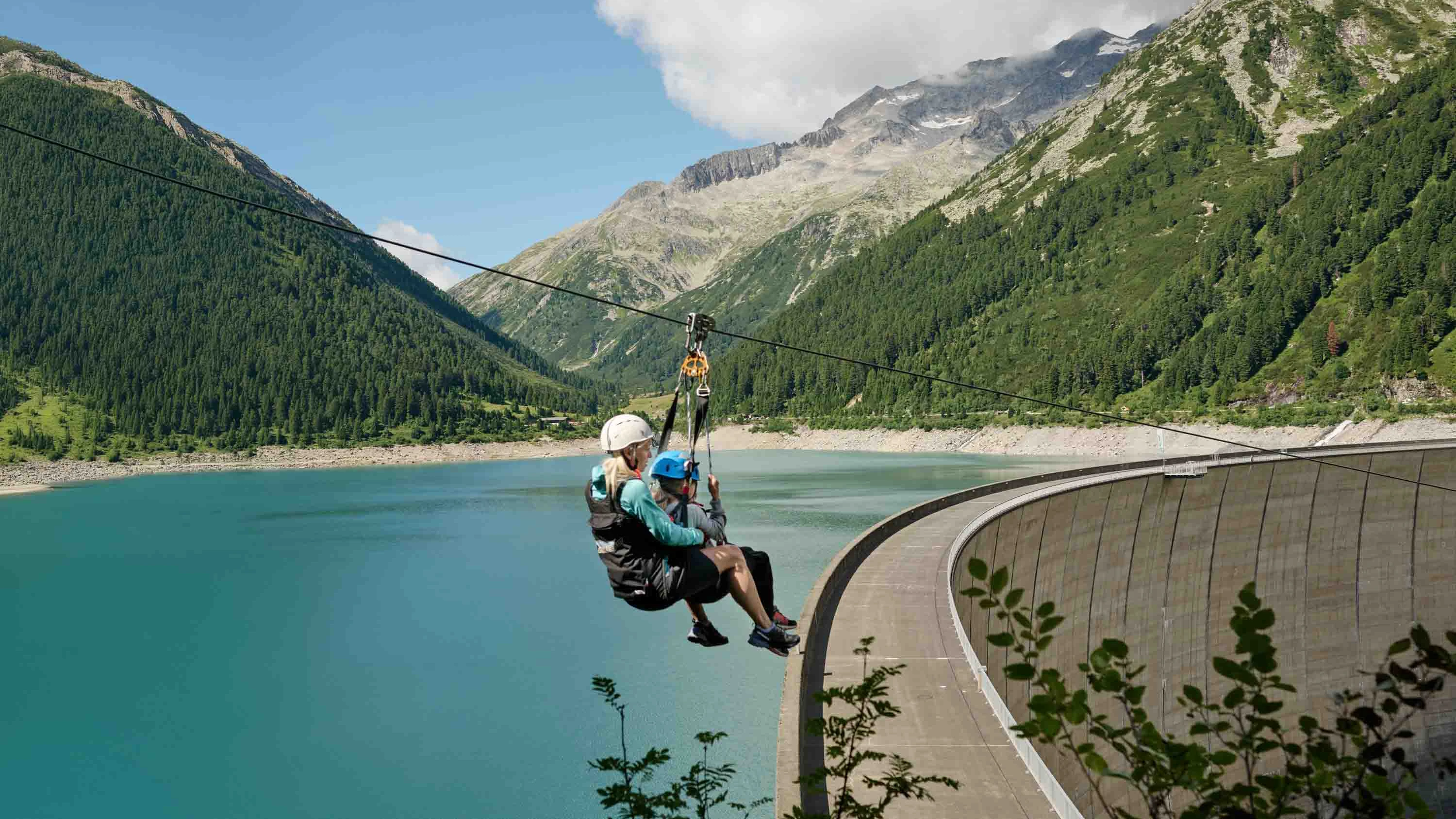 Eine Mutter mit ihrem Kind sausen den Flying Fox in Schlegeis hinab. Im Hintergrund sehen wir den See, Berge, Wälder und die Staumauer.