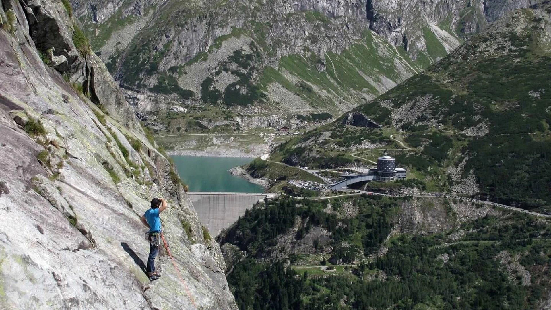 Auf der linken Seite des Bildes sehen wir einen Kletterer am Berghang stehen. Dieser blickt nach hinten rechts auf das Berghotel Malta. Zu sehen sind außerdem der Stausee und die Berge.