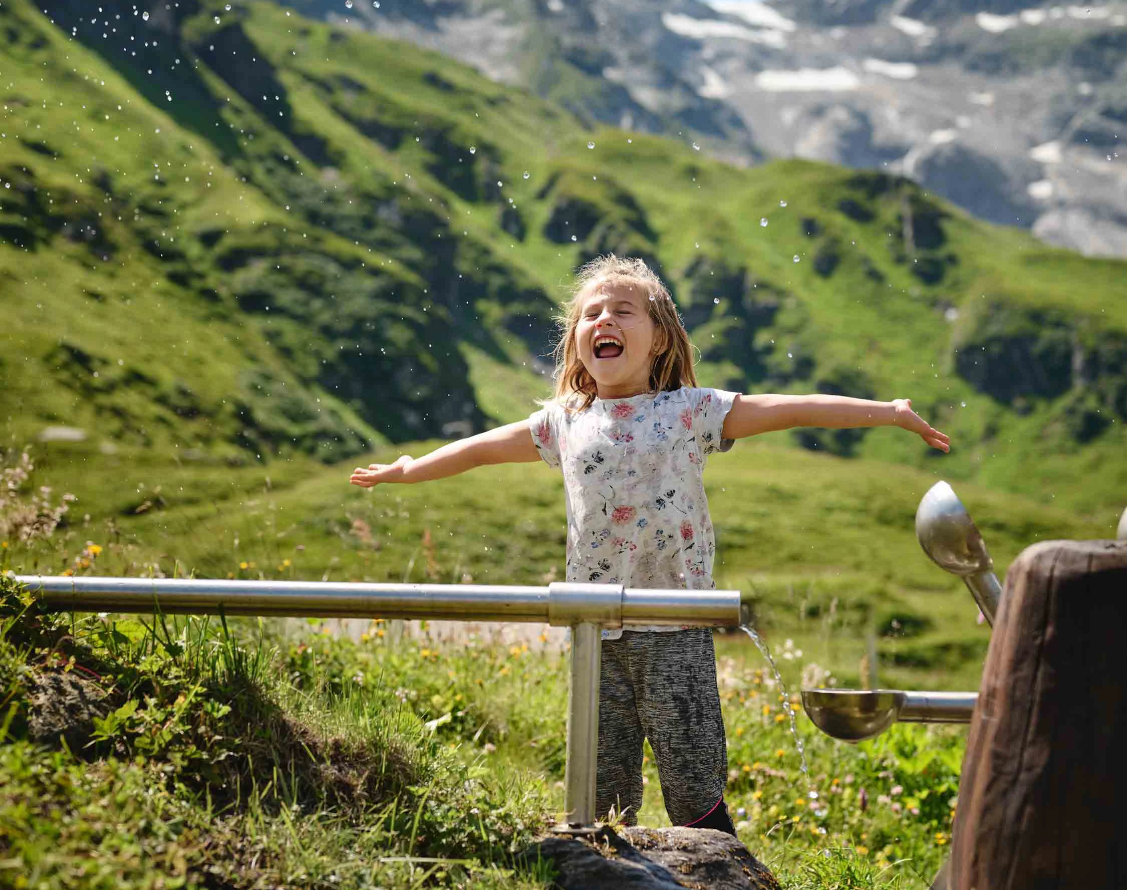 A child is standing in front of a kind of water toy, with their arms outstretched and their eyes closed. The water droplets glisten against the bright blue sky.