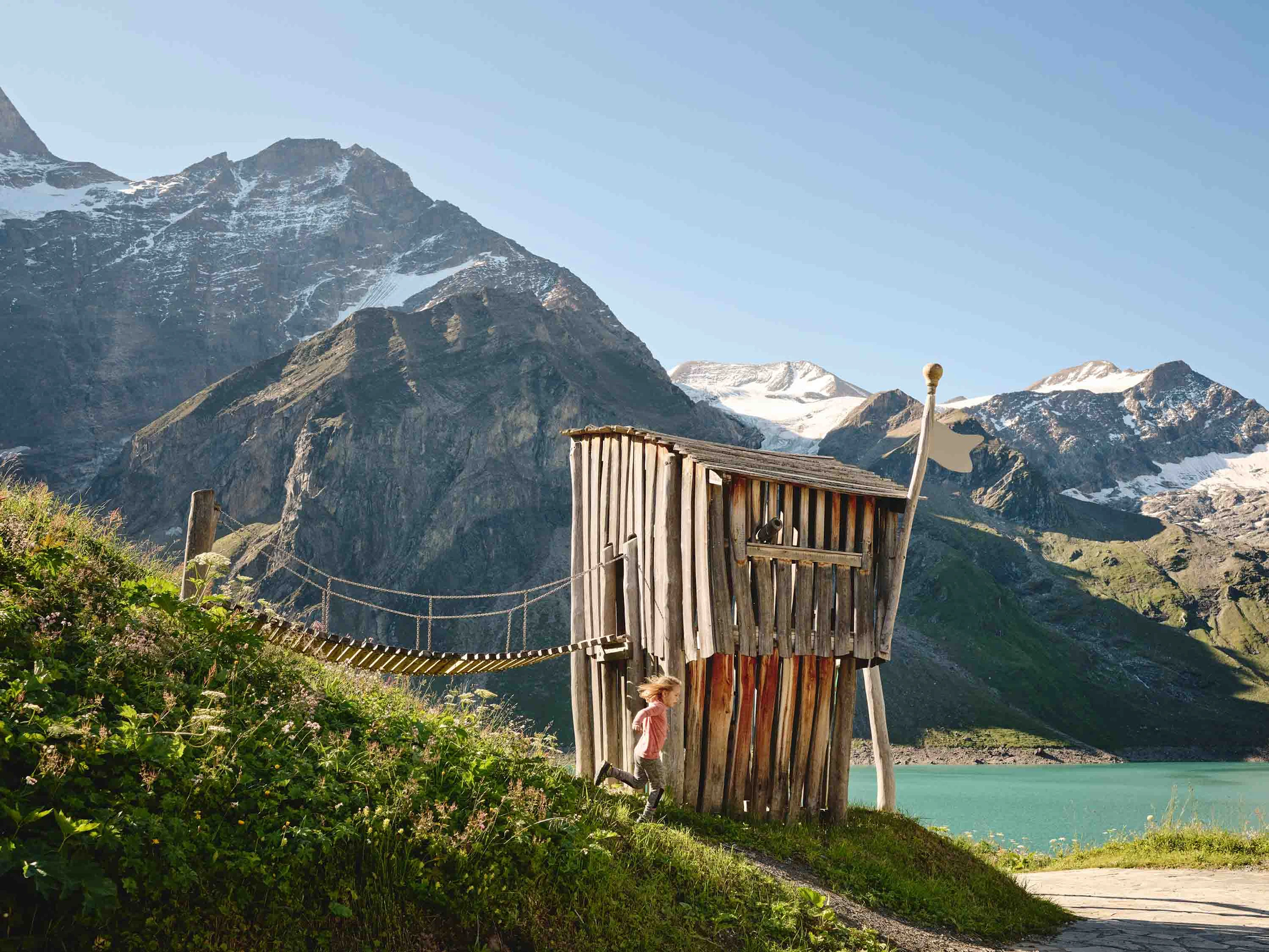 A view of the Mooserboden in Kaprun. In the foreground, we can see a child running down a slope. In the middle ground, there is a wooden hut with a suspension bridge, and in the background are the lake and the mountains.