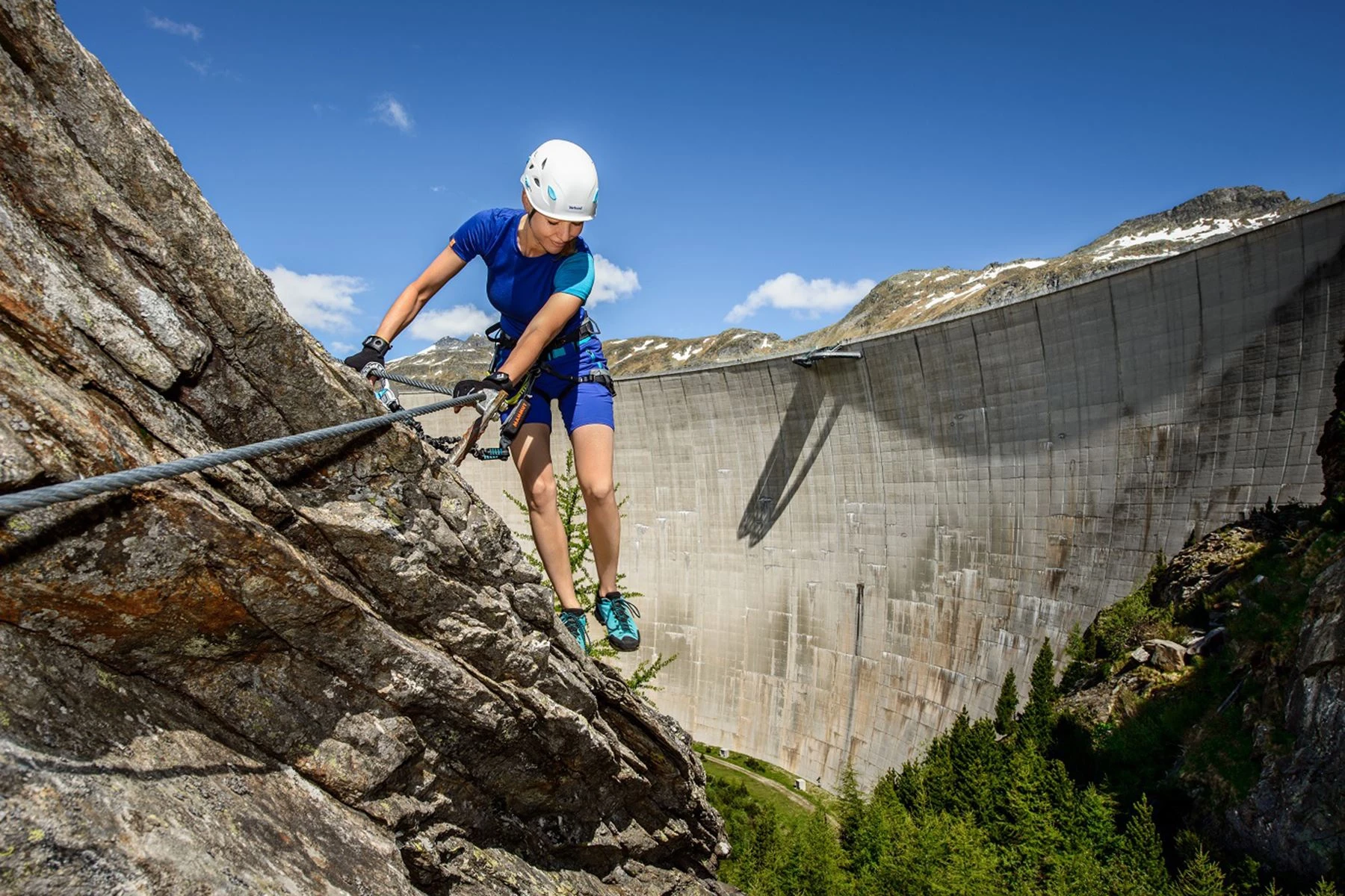 We are looking at a woman directly on the Dam High via ferrata in Malta. She is wearing dark blue summer climbing gear and a white helmet and is holding on to a rope. The large dam wall of Malta looms in the background.  The sky in the background is almost cloudless with bright sunshine.