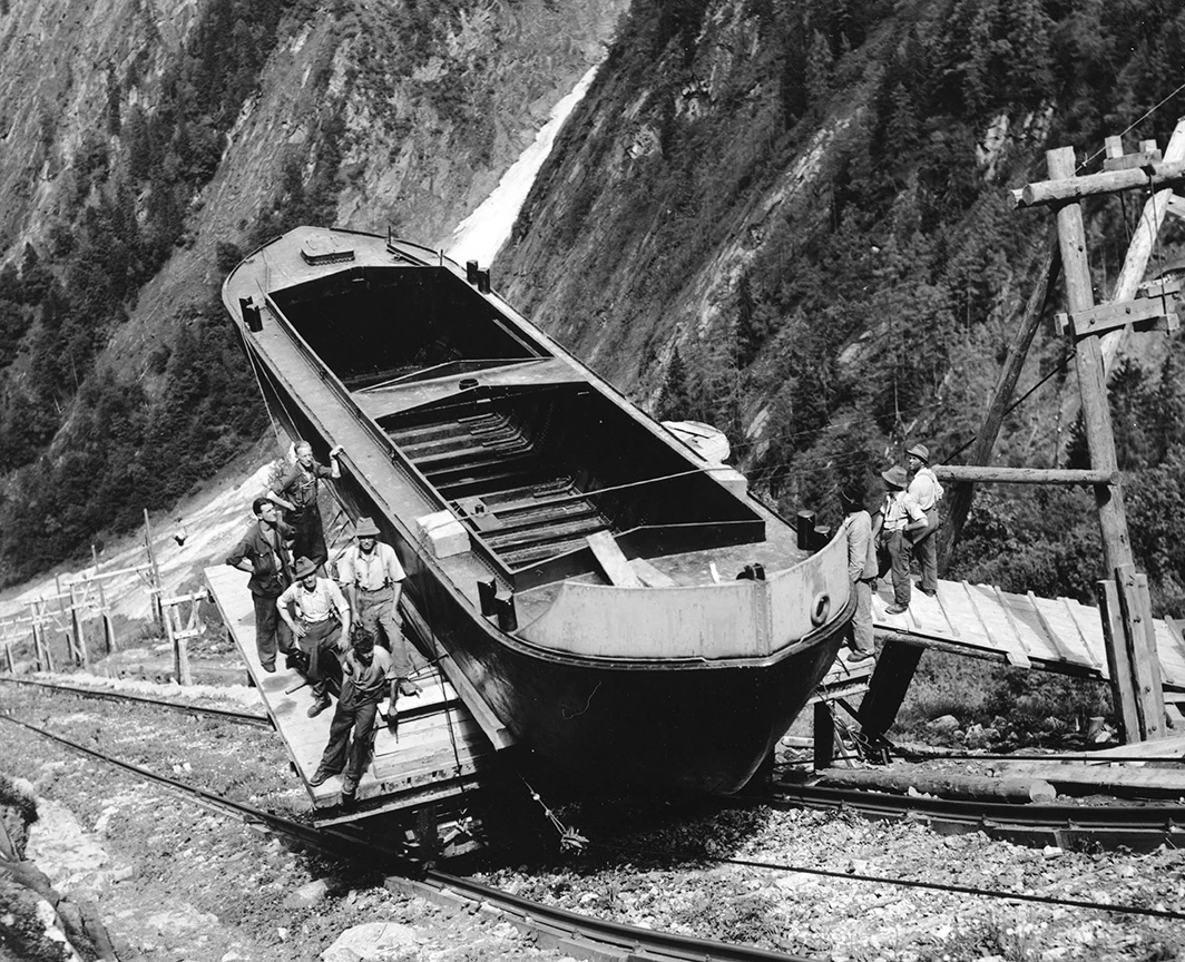 Wir sehen ein historisches Schwarz-Weiß-Bild der Erstversion des Lärchwand Schrägaufzug. Dieser besteht aus einer einfachen stabilen Holzplatte. Darauf werden einige Männer und ein Bot transportiert.