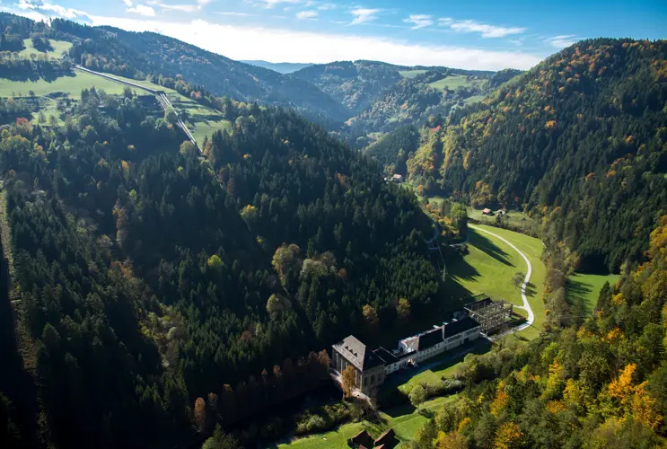 A view from above of the Arnstein power station in Styria. It is surrounded by meadows and forests. The sun is shining.