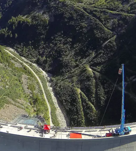 Die Flugaufnahme zeigt das Bungee-Jumping-Spektakel auf der Malta Staumauer im Sommer. Wir sehen einen großen Kran mit Korb aus dem Interessierte sich in die Tiefe stürzen können.