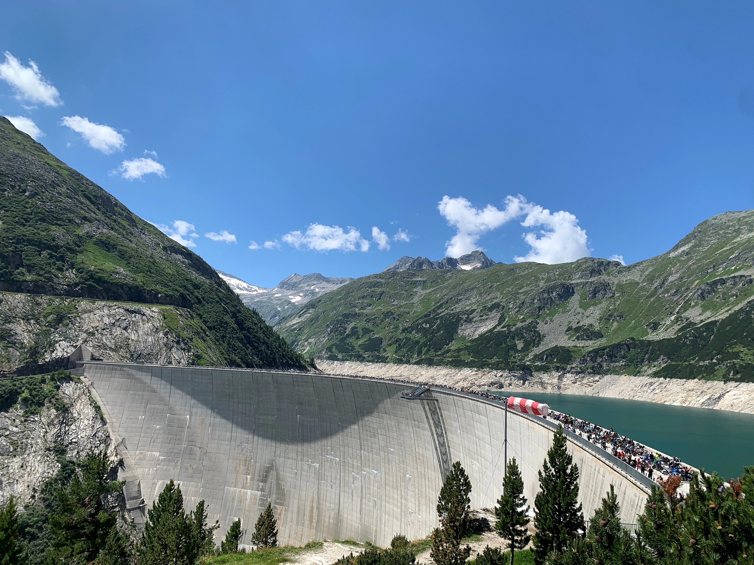 Wir blicken auf die Staumauer in Malta bei strahlenden Sonnenschein. Darauf haben sich hunderte Motorradfahrer für die Segnung ihres Bikes versammelt.