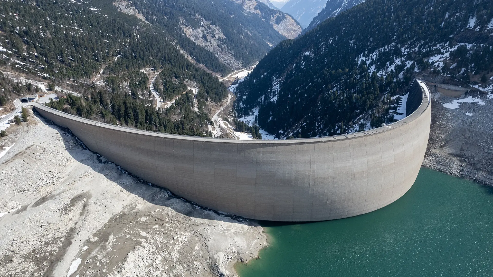 A bird's eye view of the reservoir in Schlegeis. The sun shines on a snow-covered mountain landscape.