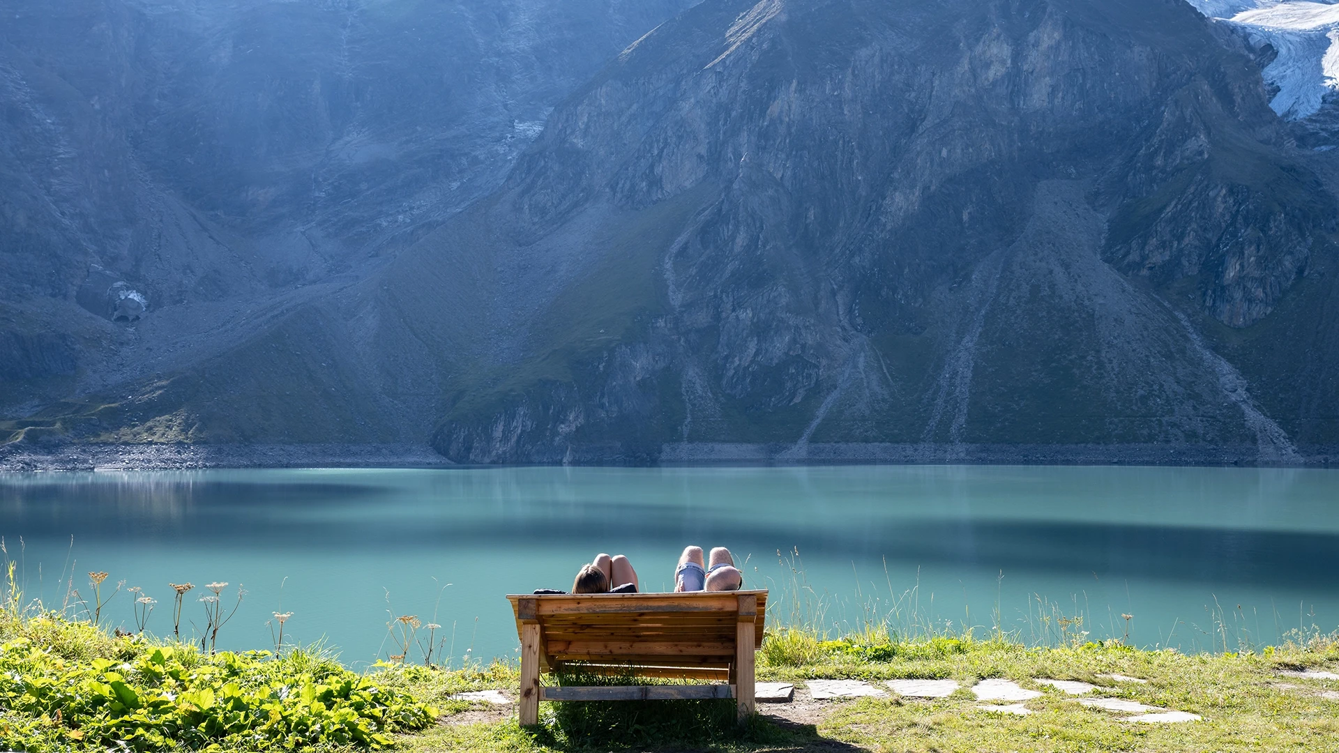 We see a summery mountain landscape, a reservoir in front of it and a sun lounger with two people on it, sunbathing in summer clothes and enjoying the mountain panorama.