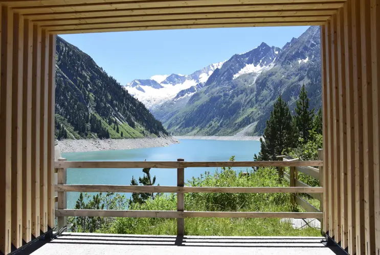 We look through a wooden construction onto the reservoir in Schlegeis. The water is a marvellous blue and the sun is shining. It's summer.