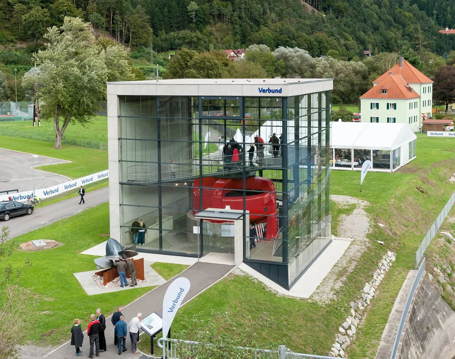 Ein Blick von ob auf die Schauturbine in Pergnegg. Diese befindet sich in einem Betongebäude mit 3 Glasfronten. Die Turbine ist rot. Zahlreiche Besucher stehen im Gebäude und außen. Die Umgebung ist sehr ländlich.
