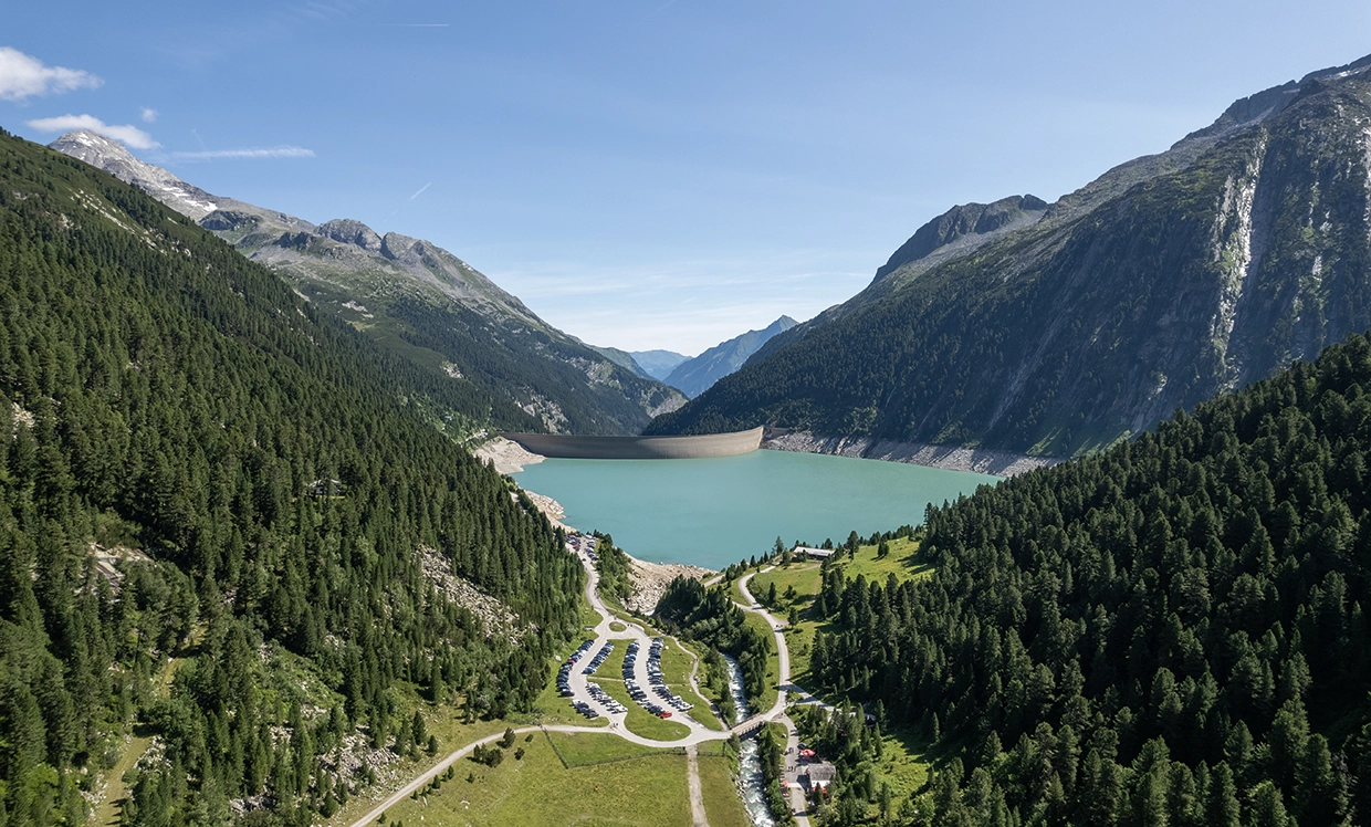 Wir blicken aus der Ferne auf den Stausee in Schlegeis. Die Wälder und Wiesen sind herrlich grün und Stausee und der Himmel strahlt unterschiedlichen Blautönen.