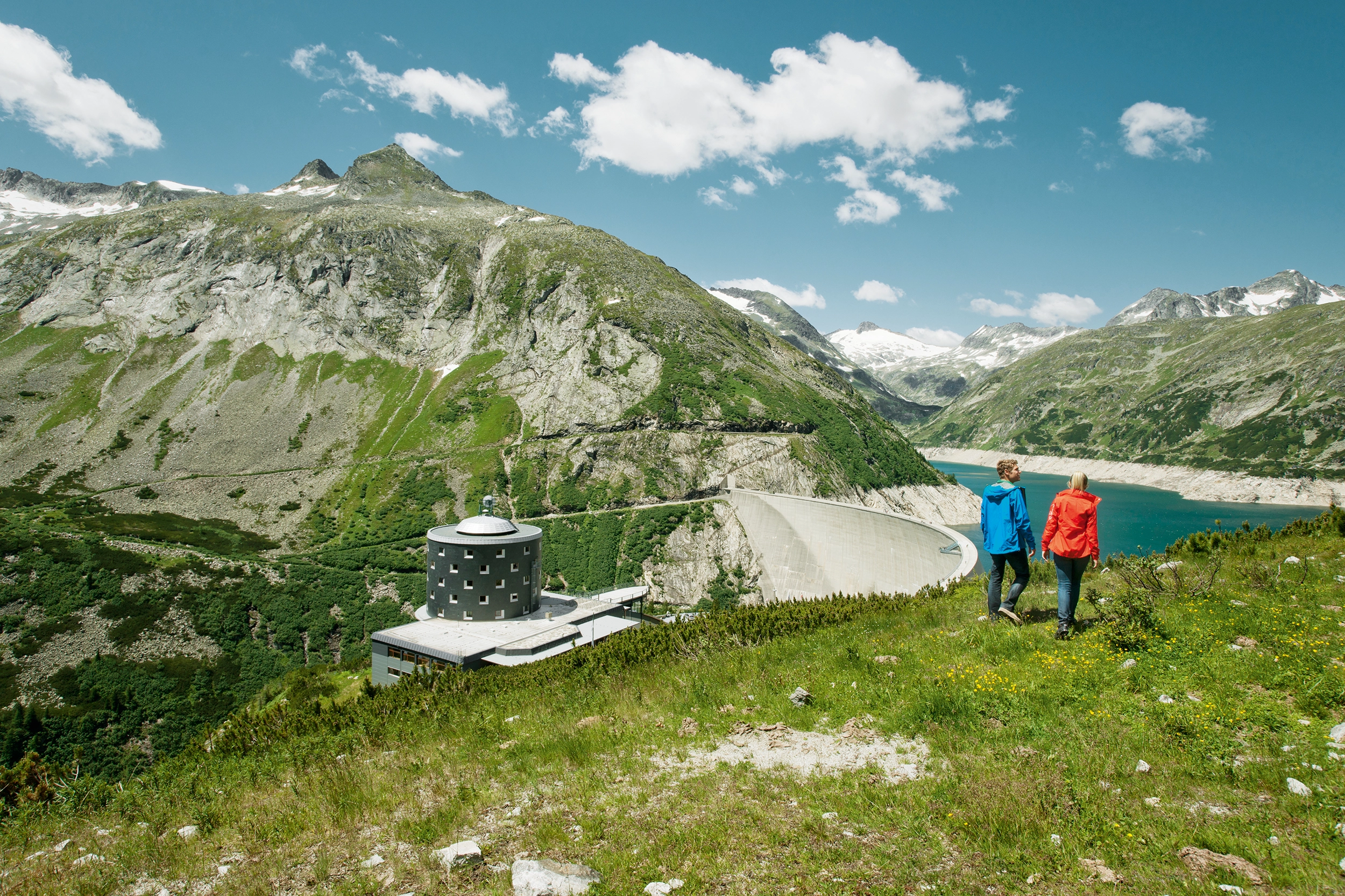 A couple stroll across the mountain meadows in Malta. In the sunshine, they have a view of the reservoir and the Malta mountain hotel, which is offset to the left.