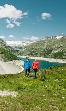 A couple stroll across the mountain meadows in Malta. In the sunshine, they have a view of the reservoir and the Malta mountain hotel, which is offset to the left.