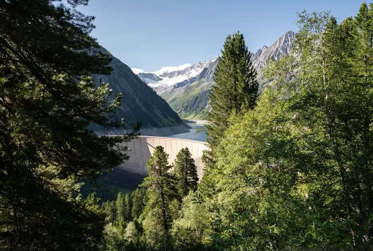 We look at the dam wall in Schlegeis from above. It is surrounded by green trees. The sun is shining.