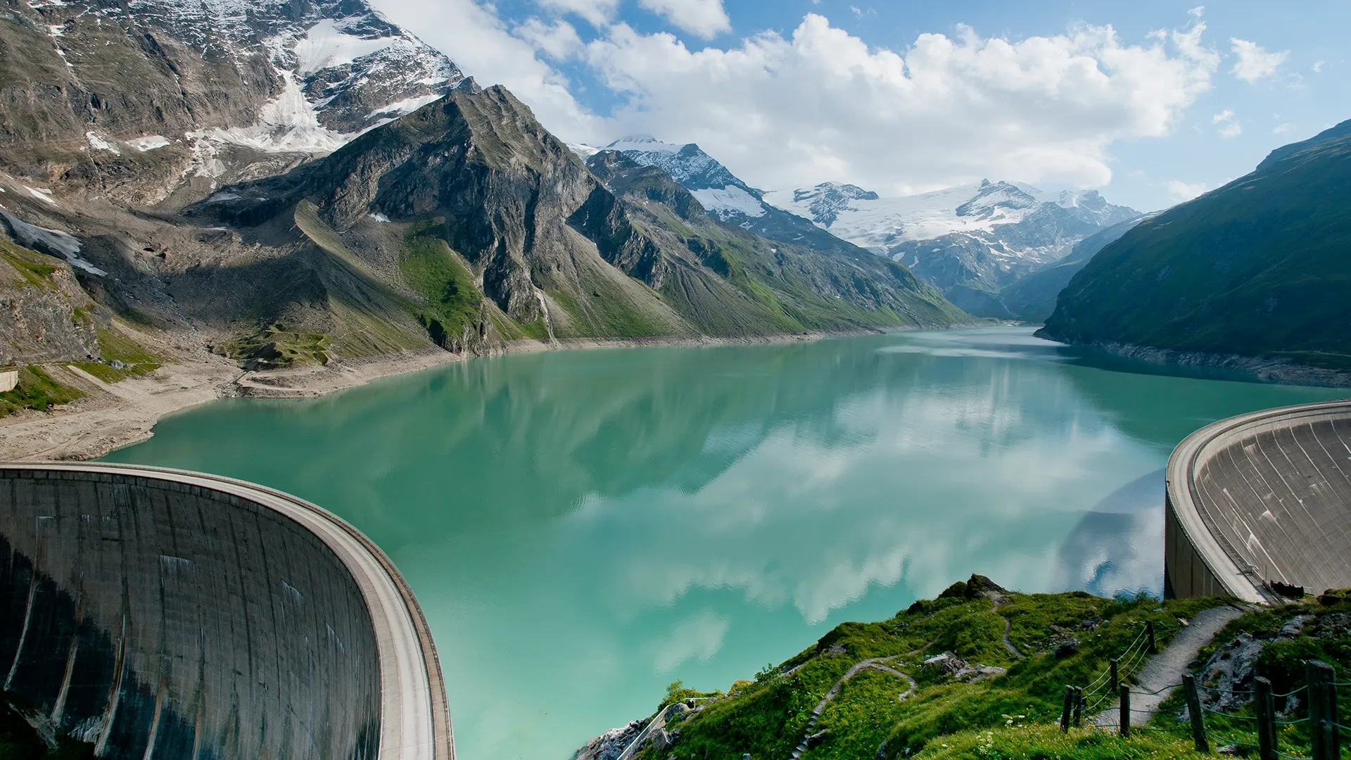 You can see a high mountain panorama in the sunshine. In front of it is a reservoir with a dam wall. The blue sky is covered with fluffy white clouds, the sun is shining and makes the reservoir water glow turquoise. The meadow banks are lush green.