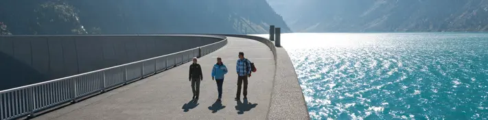 Drei Besucher spazieren über eine Staumauer. Auf der rechten Seite sehen wir den Stausee. Die Sonne scheint.