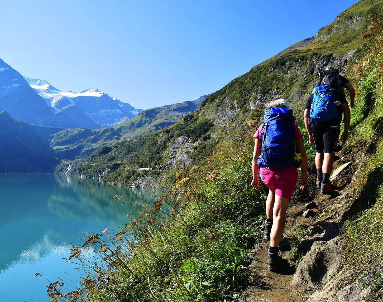 Wir blicken zwei Wandernden hinterher. Sie gehen rechts vom Stausee Mooserboden in Kaprun einen schmalen Weg empor. Im Hintergrund sehen wir die Alpen. Der Himmel ist strahlend blau.