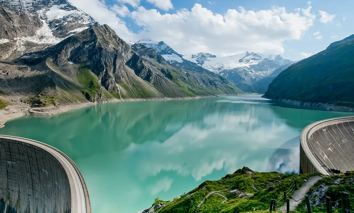 We look out over the Mooserboden reservoir in Kaprun. The clouds are reflected in the blue-turquoise water. The peaks in the background are covered in snow. The sun is shining and the meadows are green.