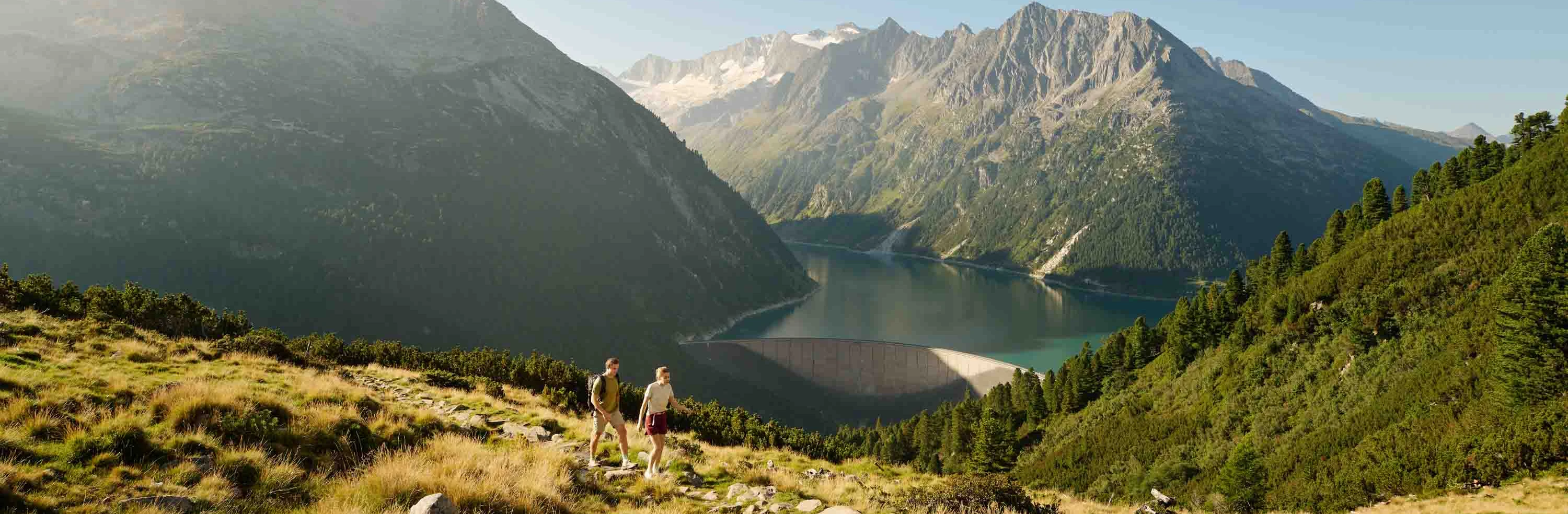 A view of the Schlegeis reservoir. In the foreground, we can see two hikers. In the background, the mountains rise up.