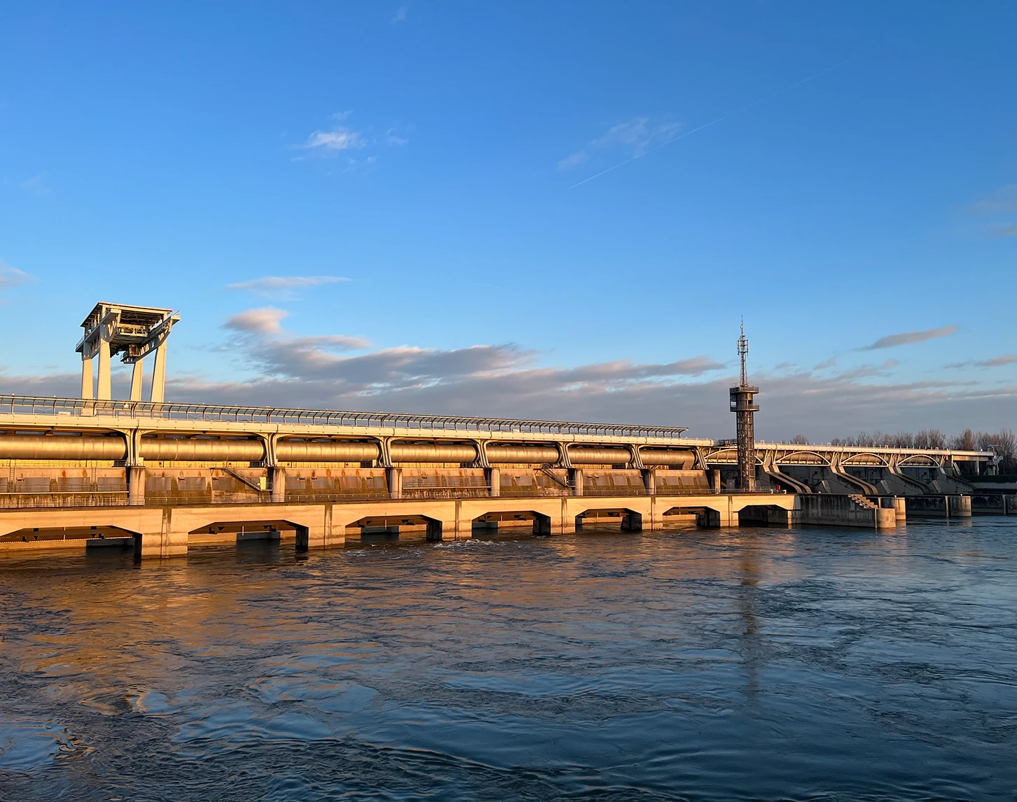 We look at the Freudenau hydroelectric power station from outside at sunset. The power station is lit up in shades of orange. The sky and the Danube are blue.