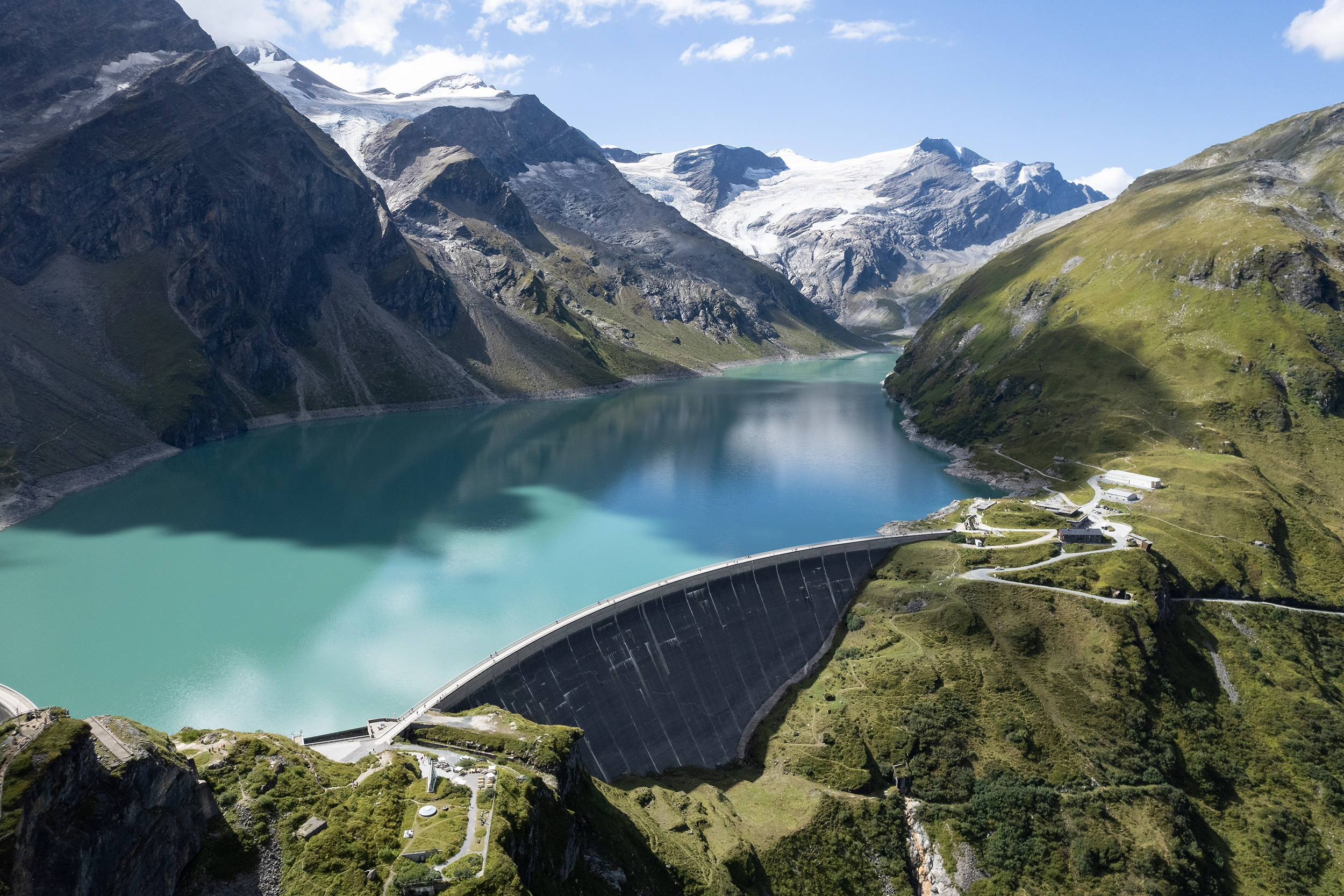 We are looking from above at the Mooserboden reservoir in Kaprun. We can see the dam wall in the foreground. The Alps tower up in the background. The sun is shining.