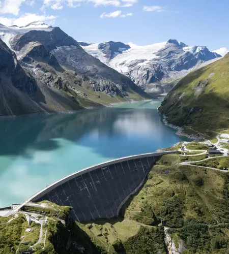 Wir blicken von oben auf den Stausee Mooserboden in Kaprun. Die Staumauer sehen wir im Vordergrund. Die Alpen türmen sich im Hintergrund auf. Die Sonne scheint.
