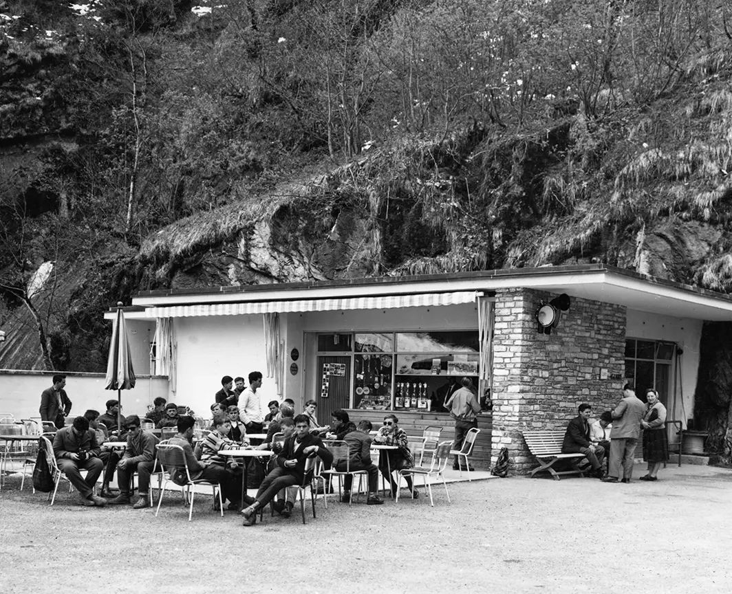 Wir sehen ein historisches Schwarz-Weiß-Bild. Zusehen ist ein Kiosk im Sommer. Davor befinden sich zahlreiche Tische und Stühle, die vollbesetzt sind.