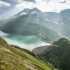 We look down on the Mooserboden reservoir in Kaprun from above. The atmosphere is spring-like. A light mist hovers over the lake. The sun's rays shine in between.