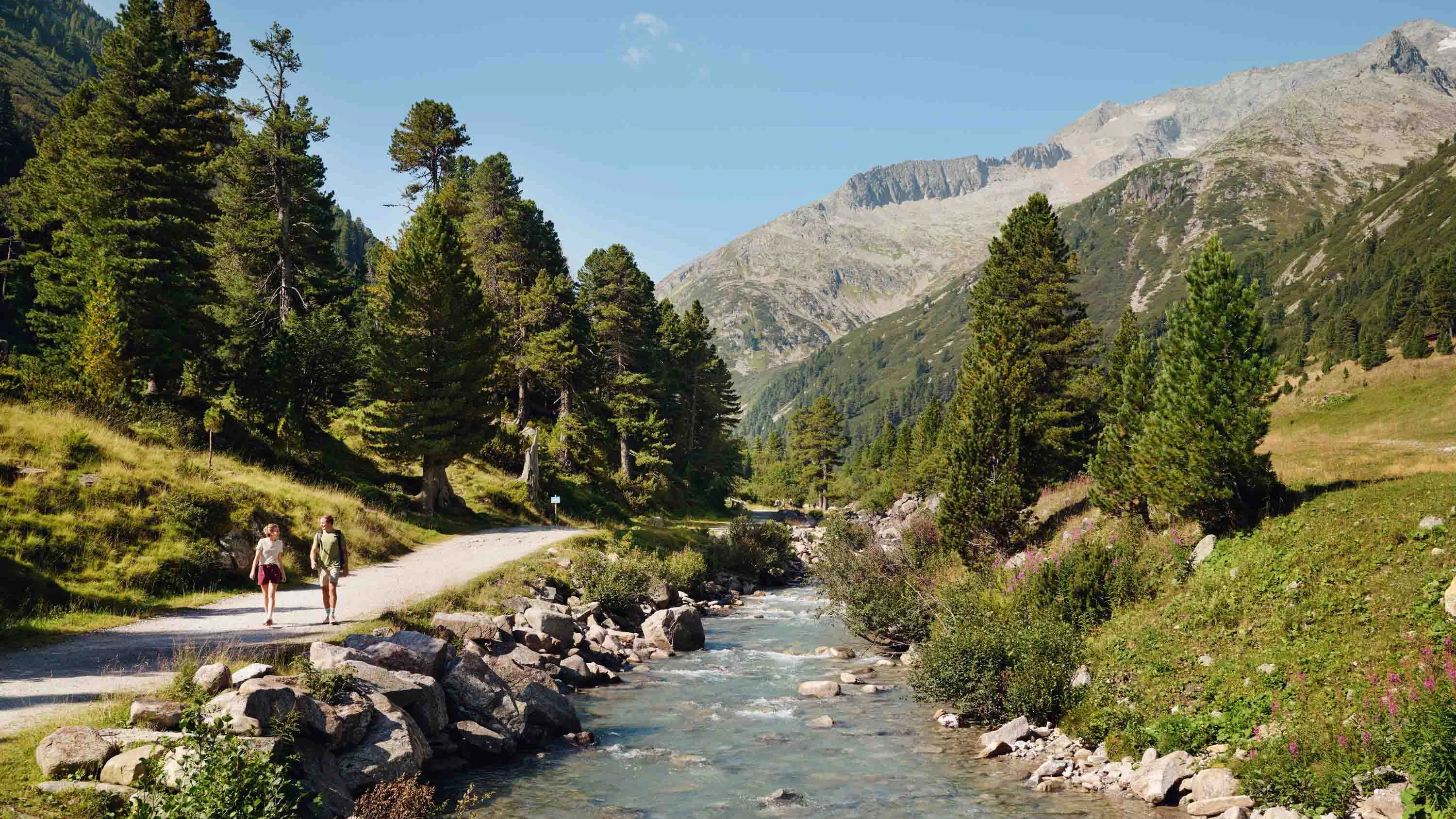 Am linken Bildrand schlängelt sich ein Wanderweg an einem Gebirgsfluss vorbei. Im Hintergrund sieht man Berge. Im Vordergrund Wiesen und Wälder.