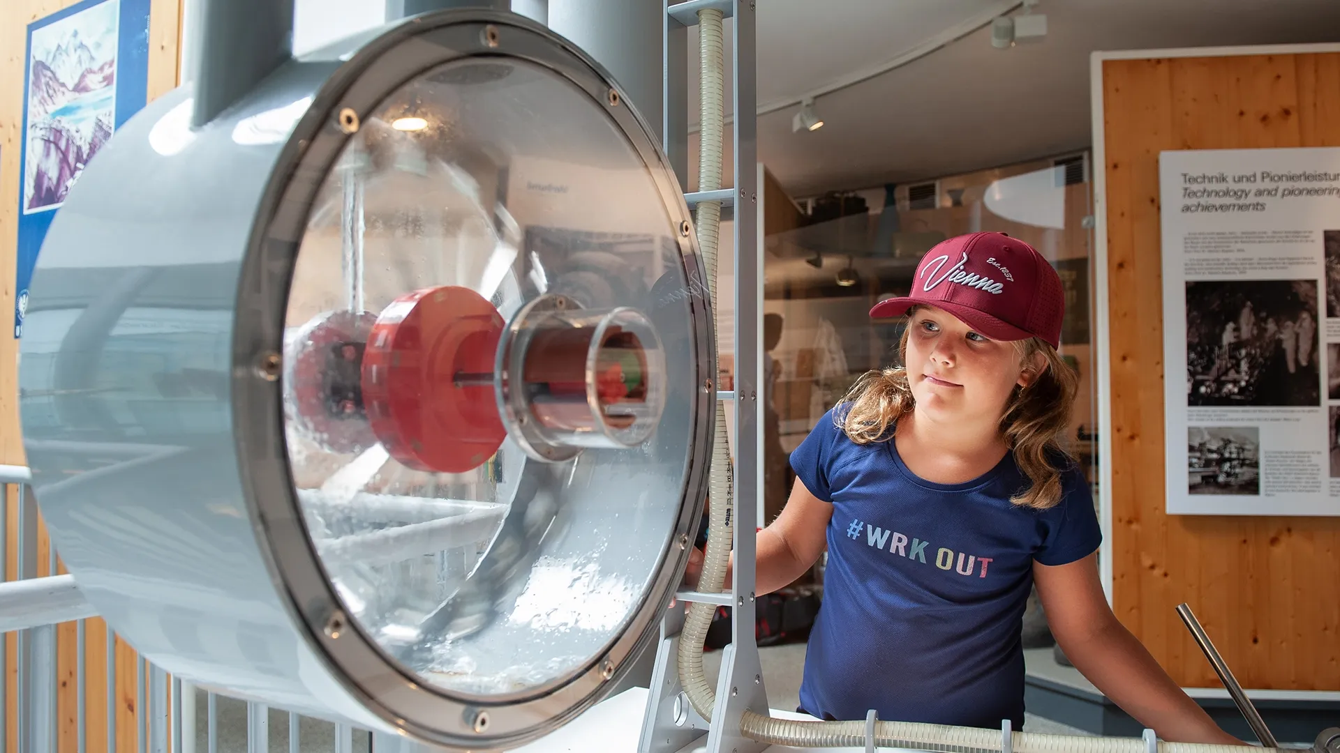 Auf der rechten Bildseite sehen wir ein kleines Mädchen. Sie trägt eine dunkelrote Cappy und ein dunkelblaues T-Shirt. Sie befindet sich in der Ausstellung der Erlebniswelt Strom in Kaprun. Sie blickt interessierte auf eine runde Apparatur mit Plexiglas.