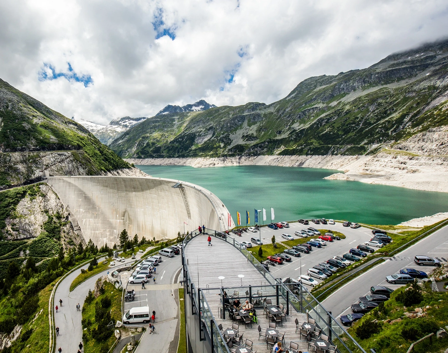 We are looking from an elevated position at the reservoir with wall in Malta. In the foreground we can see the car park and the Malta Visitor Centre. Here you can relax on the sun terrace and enjoy nature. Numerous cars and people can be seen.