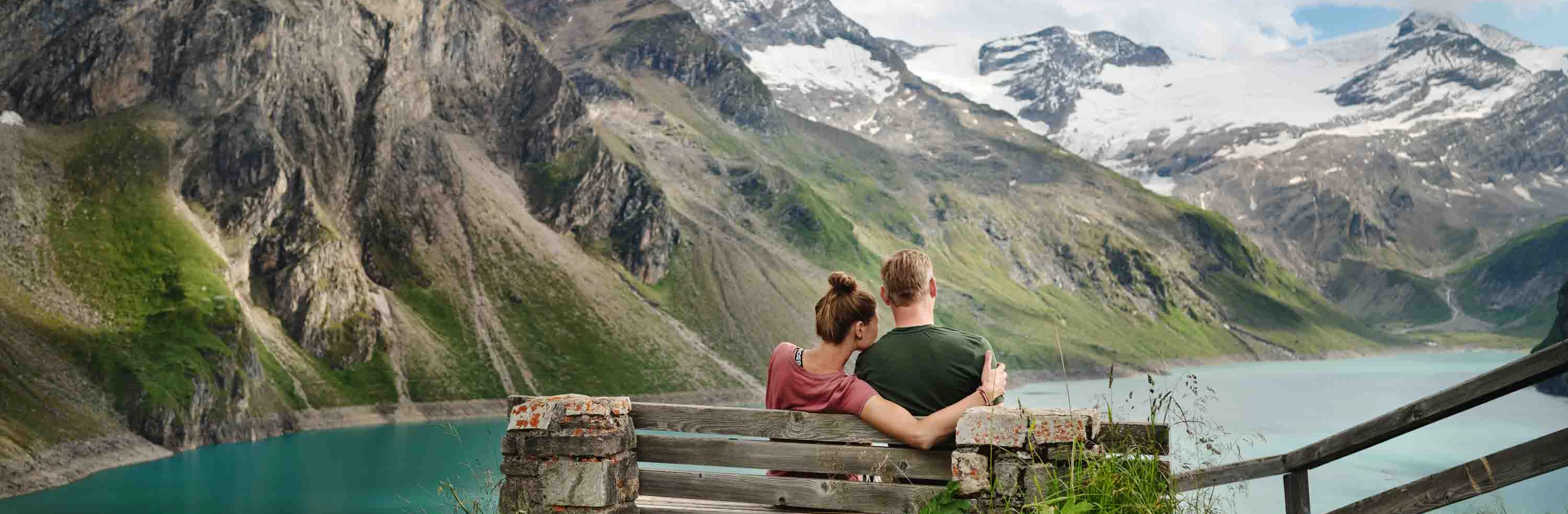 A young couple are sitting on a bench at the hilltop castle. They are looking out over the reservoir. Snow-capped mountains stretch out in the background.