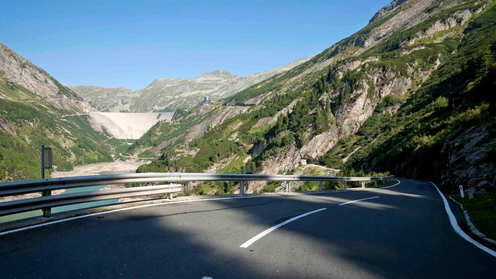 Wir blicken von der Auffahrtsstraße in Richtung Kölnbrensperre. Die Staumauer ist im Hintergrund auf der linken Bildseite zu sehen. Der Himmel ist blau und die Berg sind auf die dieser Höhe noch leicht begrünt.