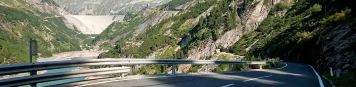 Wir blicken von der Auffahrtsstraße in Richtung Kölnbrensperre. Die Staumauer ist im Hintergrund auf der linken Bildseite zu sehen. Der Himmel ist blau und die Berg sind auf die dieser Höhe noch leicht begrünt.