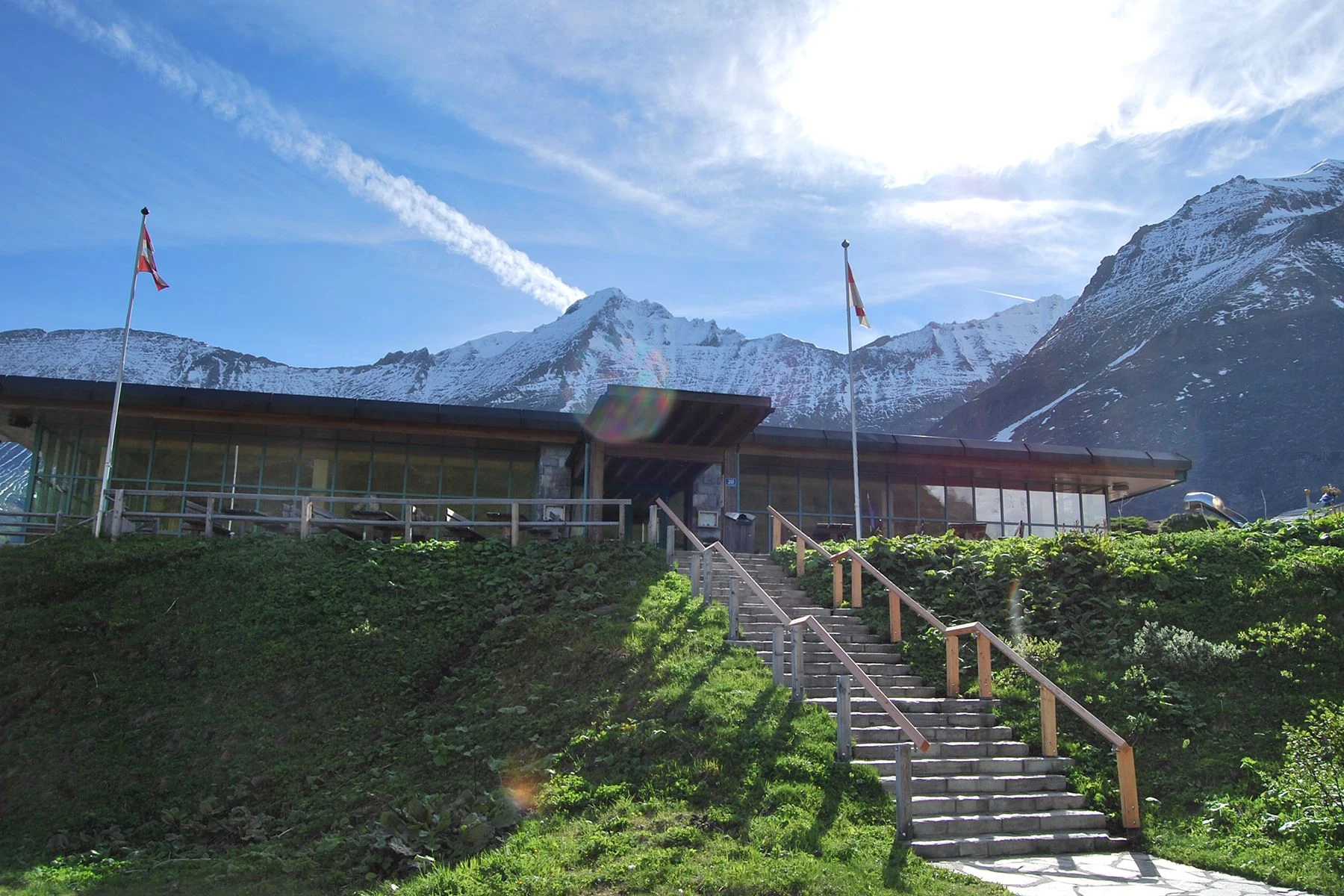 We see the entrance to the Mooserboden mountain restaurant from the outside. Stairs lead up a meadow hill to the restaurant pavilion. Two flags are waving in the wind in front of it. The high mountains can be seen behind the building. The sky is sunny with light clouds